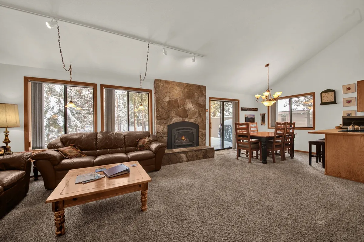 The image shows a spacious and inviting living/dining area featuring a stone fireplace, a comfortable sofa, a large dining table, and expansive windows that let in plenty of natural light. The layout encourages relaxation and group gatherings, with warm tones and a homely atmosphere ideal for families and friends.
