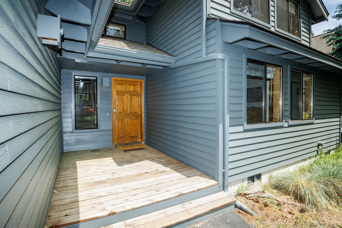 A welcoming exterior view of the home, featuring a wooden front door, entrance porch, and large windows. The lush greenery and spacious porch suggest a seamless connection between indoors and the surrounding natural environment.