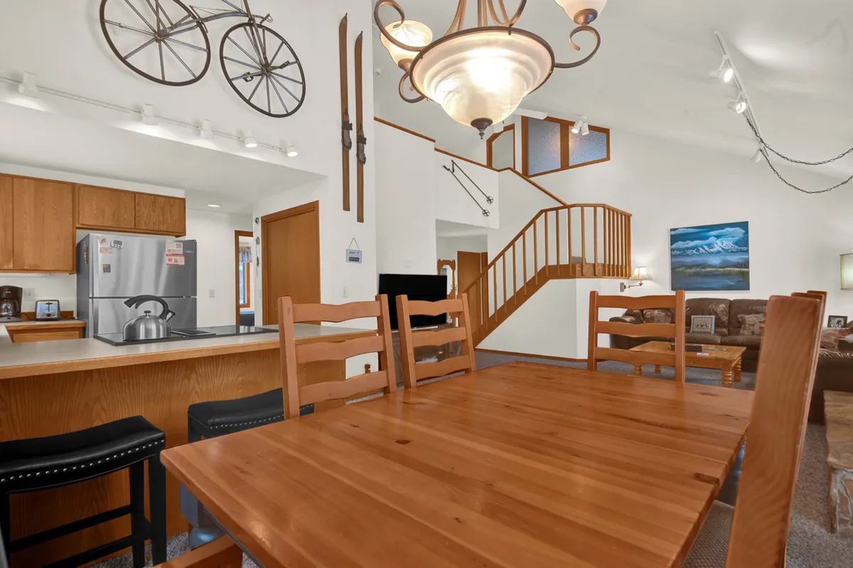 A view of the dining area with a large table under a modern chandelier, adjacent to a well-equipped kitchen and featuring a glimpse of a staircase and tasteful artwork. The scene feels both spacious and intimate, ideal for festive dinners or casual breakfasts.