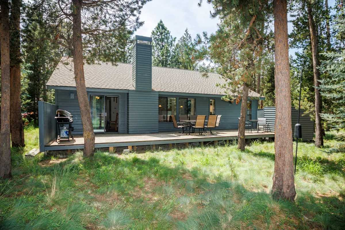 A spacious wooden deck surrounded by tall trees features comfortable outdoor seating and a grill, creating the ideal setting for al fresco dining and relaxing in nature. The chimney suggests warmth for cool evenings, and the deck looks out into the tranquil forest setting.