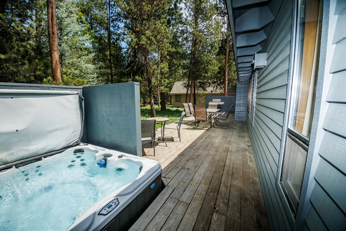 The image displays the deck area from a different perspective, highlighting the bubbling hot tub, outdoor furniture, and the surrounding forested scenery. The environment feels peaceful and invites relaxation, perfectly aligning with the narrative of wildlife views and starlit evenings.