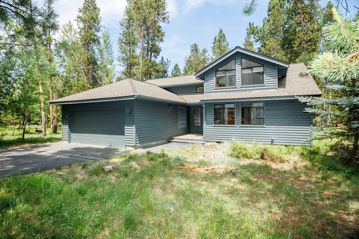 The home's welcoming facade framed by mature trees, with a driveway hinting at plenty of space for arrivals and outdoor play. The tranquil, green setting invites families to enjoy the peaceful Sunriver environment and the sense of coming home.